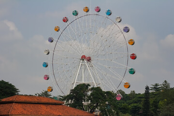 Fototapeta premium A large Ferris wheel with colorful cabins set against a cloudy blue sky and trees.