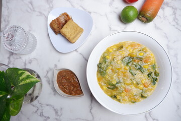 A traditional Indonesian meal featuring Bubur Manado or porridge with vegetables and salted fish on a classic plate, accompanied by fried tofu and a glass of water. A top view of traditional Bubur Man