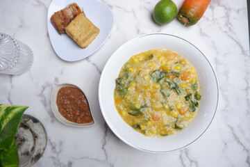 A traditional Indonesian meal featuring Bubur Manado or porridge with vegetables and salted fish on a classic plate, accompanied by fried tofu and a glass of water. A top view of traditional Bubur Man