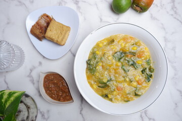 A traditional Indonesian meal featuring Bubur Manado or porridge with vegetables and salted fish on a classic plate, accompanied by fried tofu and a glass of water. A top view of traditional Bubur Man