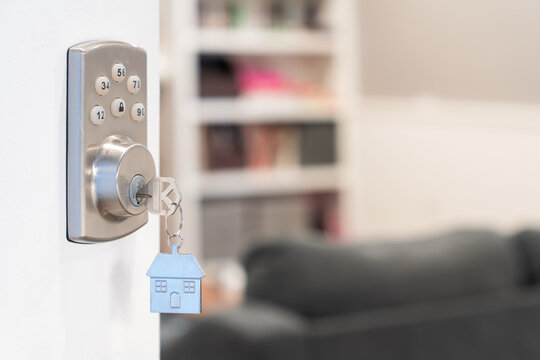 House key with a silver house shaped keychain hanging from a smart lock in a wooden door, open into living room symbolizing welcome or new ownership