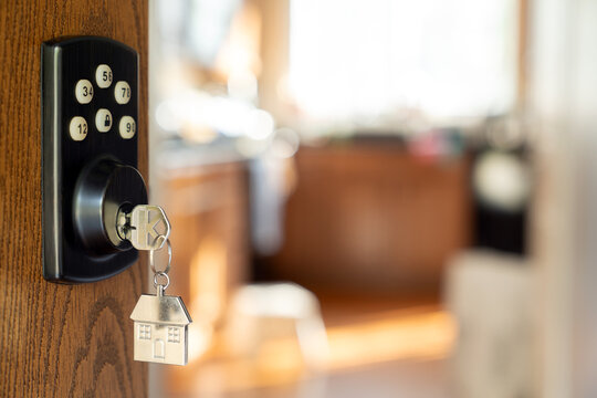 House key with a silver house shaped keychain hanging from a smart lock in a wooden door, open into living room symbolizing welcome or new ownership