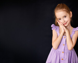 Adorable girl in lavender dress poses against dark background. 
Smiling child portrait radiates sweetness and gentle confidence.
Minimalist studio shot captures elegance, innocence, and charm.

