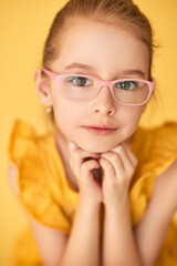 Smart girl in pink glasses poses in yellow dress. 
Closeup portrait shows innocence, confidence, and style.
Soft natural light highlights beauty and gentle expression.
