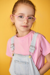 Stylish girl in pink glasses and denim overalls poses confidently. 
Child portrait on yellow background shows modern casual fashion.
Soft light highlights innocence, intelligence, and gentle charm.