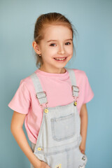 Smiling girl in pink t-shirt and denim overalls poses on blue background.
Cheerful child portrait radiates happiness and confidence.
Bright studio image captures playful style and innocence.
