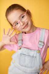 Smiling girl in pink glasses and denim overalls waves hello.
Cheerful child portrait on yellow background shows playful energy.
Bright studio photo captures happiness, style, and confidence.
