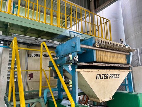 A large Filter Press machine in a wastewater treatment facility, clearly showing the dewatering mechanism and collection hopper, with yellow railings and a Sludge Thickener sign in the background.