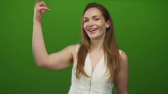 Young caucasian woman blows a kiss gesture with her fingers and smiles in a green studio; affection.