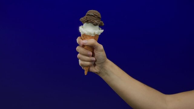 Man holding an ice cream cone against a blue background, showcasing vanilla and chocolate scoops, with a focus on the arm's texture and color contrast.