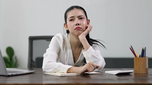 Exhausted Asian businesswoman experiencing workplace burnout, sitting at desk with laptop and stationery, mental fatigue and professional disengagement.