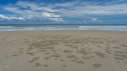 A pattern of sand bubbler crabs balls on a tropical beach. The waves of the turquoise ocean are foaming at the shore. Blue sky, clouds. Malaysia. Borneo. Kota Kinabalu.