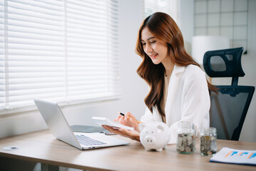 Asian woman saving for retirement Saving money through a piggy bank and taking notes in modern...