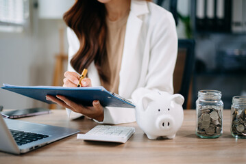 Asian woman saving for retirement Saving money through a piggy bank and taking notes in modern...