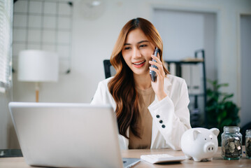 Asian woman smiling while managing finances at desk with laptop, clipboard, and coins.