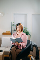 Young woman sits comfortably on a sofa, reading a notebook while holding a coffee cup.