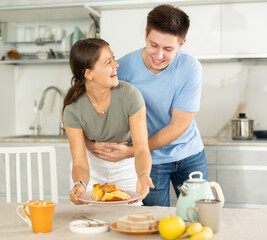 Cheerful young woman holding a plate with pie and her husband embracing her behind in the kitchen