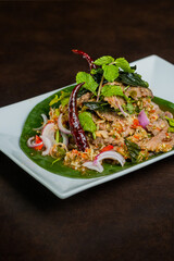 A delectable Thai dish artfully presented, showcasing vibrant ingredients and culinary expertise. The dish is placed on a white square plate, which is set atop a wooden surface