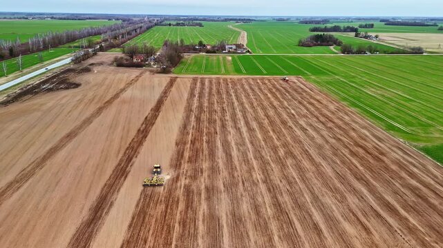 Drone shot captures two agricultural tractors with implements working simultaneously on a vast brown tilled field adjacent to vibrant green fields and scattered farm buildings during spring.
