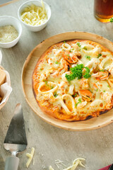 A delicious seafood pizza on a wooden plate, ready to be enjoyed. Cheese and other ingredients sit nearby.
