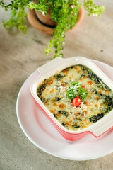 Delicious baked spinach with melted cheese, served in a square dish. Garnished with a cherry tomato and parsley on a white plate.