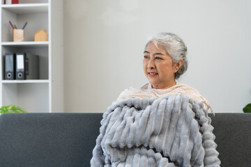 Senior Asian woman relaxing on a sofa, covered with a soft blanket, looking away with a gentle smile