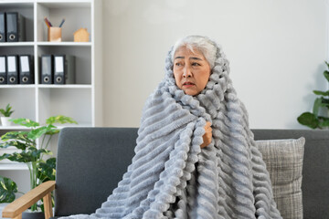 Asian senior woman feeling cold, shivering, and suffering from flu symptoms or fever while wrapped...