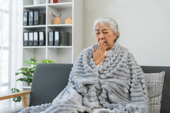 Asian elderly woman feeling sick and unwell, sitting wrapped in a blanket on a sofa at home, coughing and covering mouth