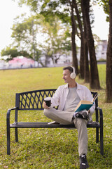 Fototapeta premium Man enjoy a leisurely moment on a park bench, listening to music with headphones, holding a takeaway coffee cup, and reading a book outdoors in a green natural setting