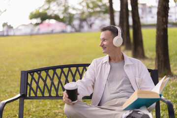 Fototapeta premium Mature man enjoying leisure time on a park bench, listening to music with headphones, holding a coffee cup, and reading a book in a serene outdoor setting