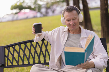 Senior man on a park bench enjoying a quiet afternoon: reading a book, sipping coffee and listening to music with headphones, relaxed and content in nature
