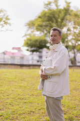 Obraz premium Senior man standing outdoors in a park, smiling and holding a modern digital tablet while wearing wireless headphones around his neck, reflecting a tech-savvy lifestyle