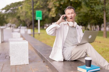 Mature man connecting remotely, multitasking with a laptop while having a surprised expression during a phone call, enjoying a coffee outdoors in a green park