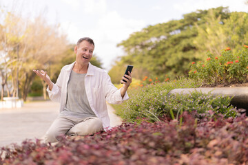 Mature man crouched in a park, holding a smartphone, looking surprised and gesturing during a video call, enjoying communication outdoors surrounded by nature