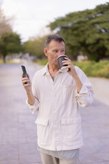 Mature man walking outdoors in a park setting, casually dressed, enjoying a takeaway coffee while holding his smartphone, representing a relaxed city lifestyle