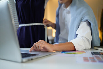 A stylishly dressed male designer creates a new collection of clothes, measures the parameters of clothes with a measuring tape and writes them down on laptop while standing in his studio.