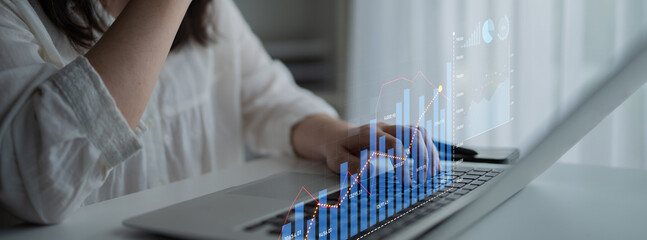 A focused woman works on her laptop in a sleek home office, analyzing financial data displayed with...