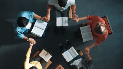 Top down view of diverse people holding hand and sitting in circle with bible book on lap. Aerial...