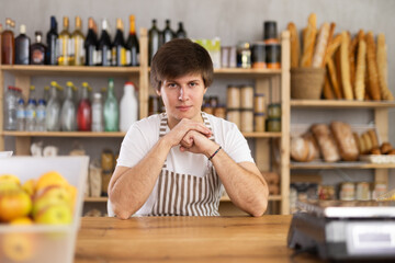 Portrait of confident young salesperson in apron sitting at counter in supermarket