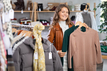 Positive young female customer reviewing winter dresses during shopping in clothing store
