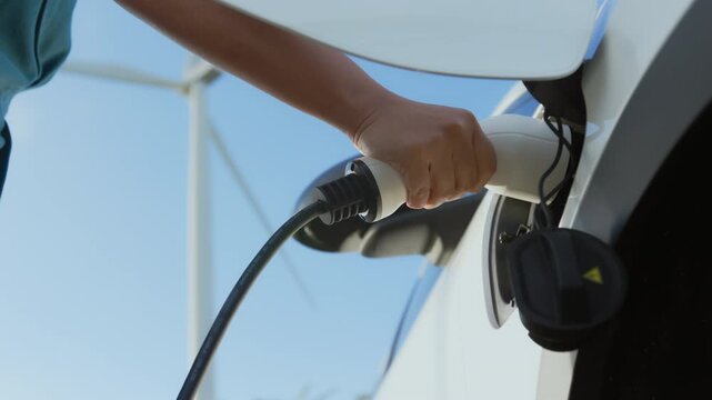 Hand plugging electric vehicle charging cable into car socket with wind turbine background, clean energy