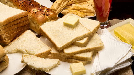 A high-quality close-up of assorted freshly baked breads arranged neatly with butter cubes, napkins, and a glass of juice on a breakfast table. The image showcases soft white bread slices