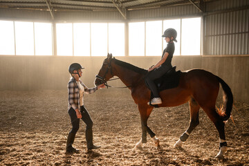 Riding instructor teaches teenage girl wearing riding helmet how to ride horse in indoor arena. Mother and daughter learning to ride. Riding school, hobby.