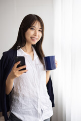 Young Woman Using Smartphone While Holding a Mug by the Window
