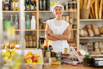 Woman salesman in white hat with set of meat, cheese, eggs and champagne behind counter in grocery...