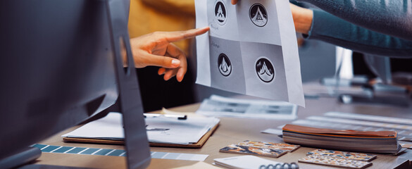 Two creative professionals engage in a collaborative discussion about logo designs. They review printed graphics and share ideas in a bright and modern office environment. SACTR