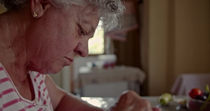 A close-up captures an elderly woman with thoughtful expression as she slowly eats a meal with a spoon, highlighting the quiet dignity and routine of senior daily life at home.