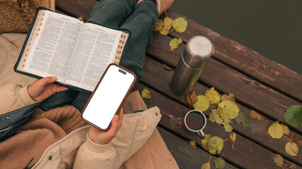 Person reading the Bible and using a smartphone outdoors on a wooden dock
