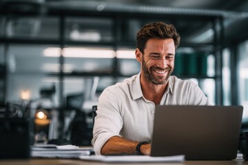 Fototapeta premium Confident middle-aged man smiling while working on a laptop in a bright modern office environment, suggesting productivity and success.