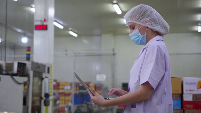 Worker woman inspector working quality control in food processing factory, woman in uniform and mask using laptop for monitoring hygiene and production, female examining quality and supply chain.